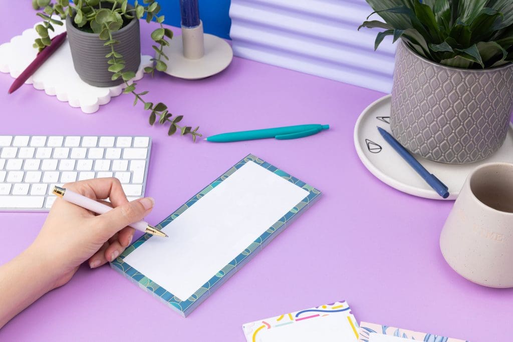 Deco delights list pad - teal gradient background with golden yellow deco motif - is on a lilac desk with pens, pot plants and white keyboard around it. There is a left hand on the left of the image holding a pen to write on the pad.