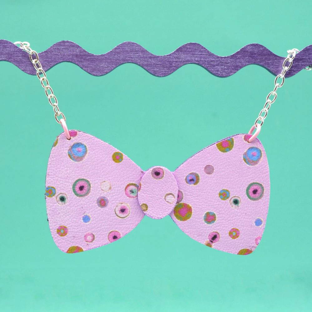 A playful bow necklace made from pale pink faux leather covered in a fun watercolour inspired abstract dot print. Photographed against a bright spearmint background