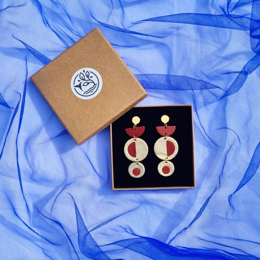 Handcrafted geometric dangly earrings with brick red inlayed in metallic gold leather, in a kraft jewellery box on a royal blue mesh fabric background.