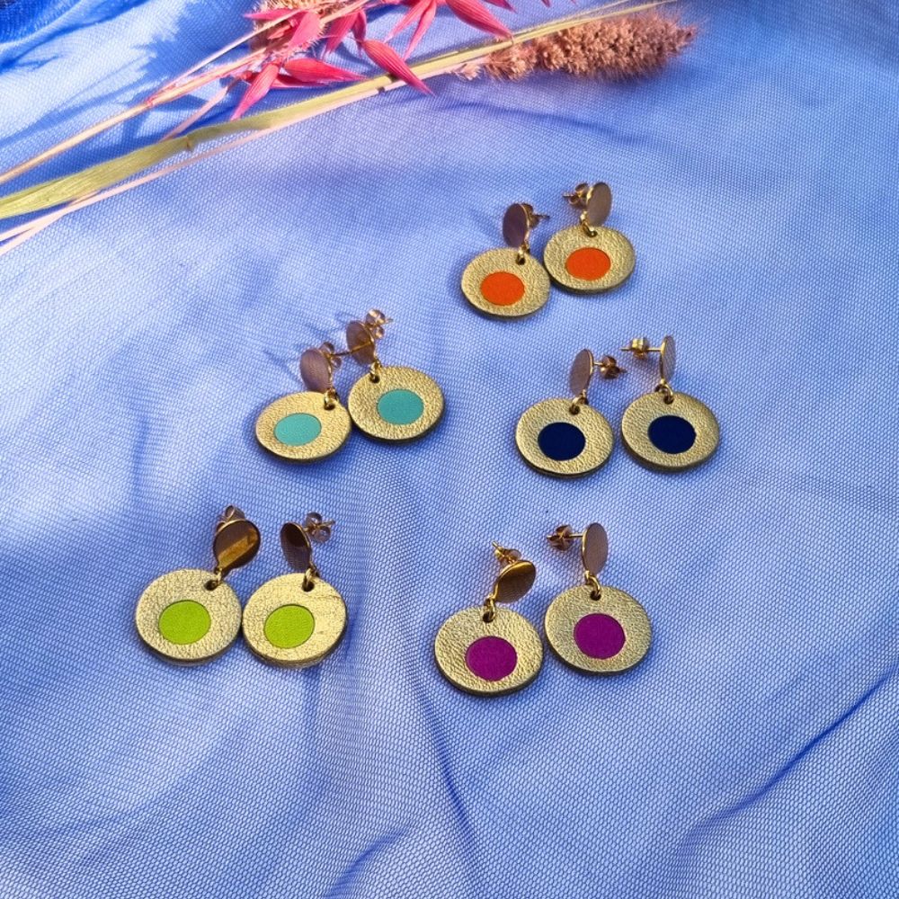 Five pairs of circular leather drop earrings in gold, featuring colourful centre inlays in orange, mint, navy, lime, and magenta, arranged on blue mesh fabric.